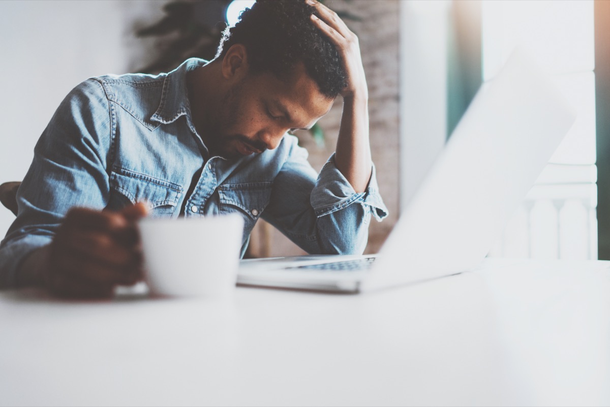 Tired young African man using laptop while sitting at the table on a sunny morning.Concept of people working hard home