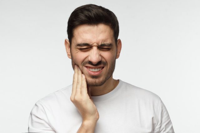 man isolated on gray background touching his face and closing eyes with expression of horrible suffer from health problem and aching tooth, showing dissatisfaction