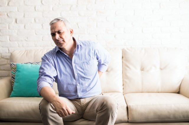 Homme mûr aux cheveux gris ayant mal au dos assis sur un canapé à la maison