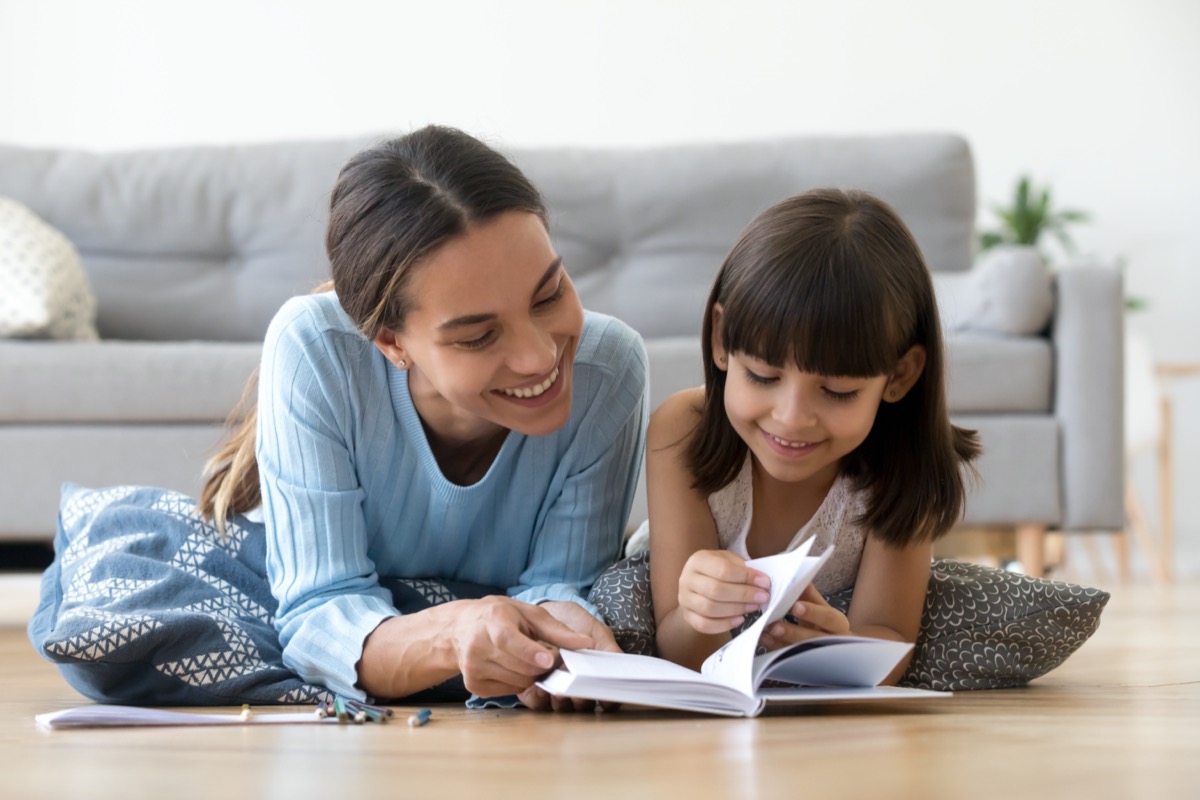 Caring smiling mother reading book with little kid girl lying on warm floor at home, mom or baby sitter playing having fun telling fairy tale to child daughter