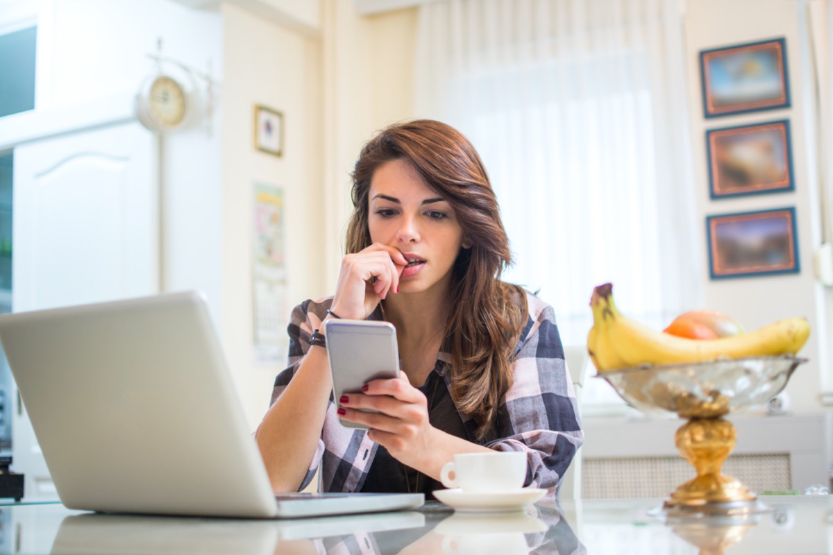 Young nervous woman looking at smartphone and biting her fingernails at home.