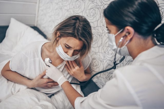 Ärztliche Krankenschwester in schützender Gesichtsmaske, die mit einem Stethoskop atmet und Coronavirus (COVID-19) vermutet.