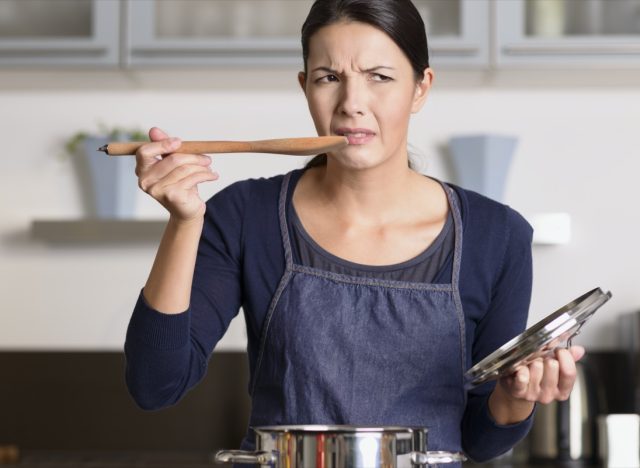 female cook standing at the hob in her apron tasting her food in the saucepan with a grimace as she finds it distasteful and unpalatable
