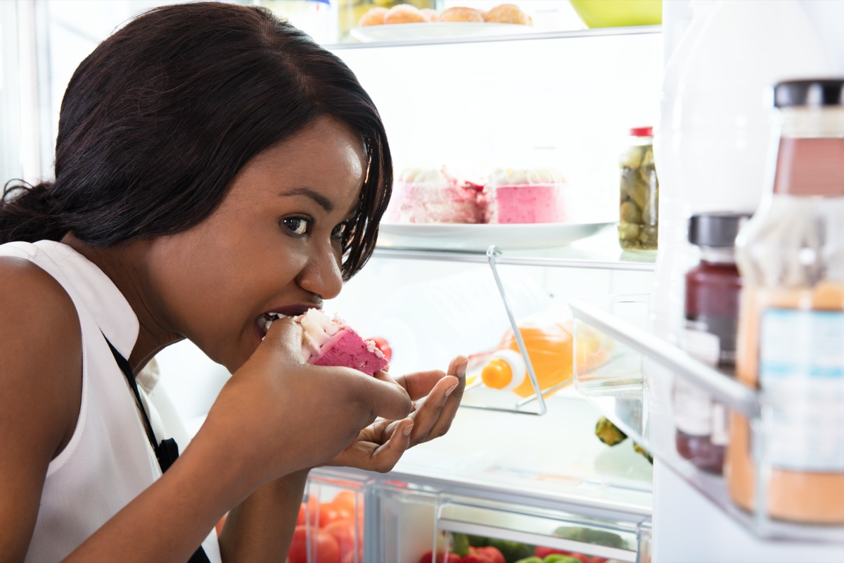 African Woman Eating Slice Of Cake Near Open Refrigerator