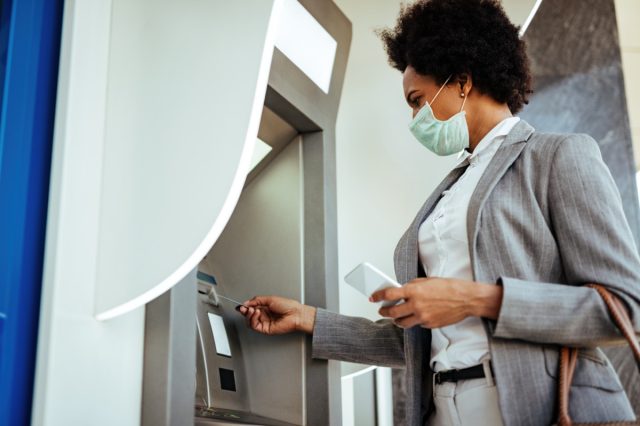 Low angle view of African American businesswoman inserting credit card and withdrawing cash at ATM while wearing protective mask on her face.