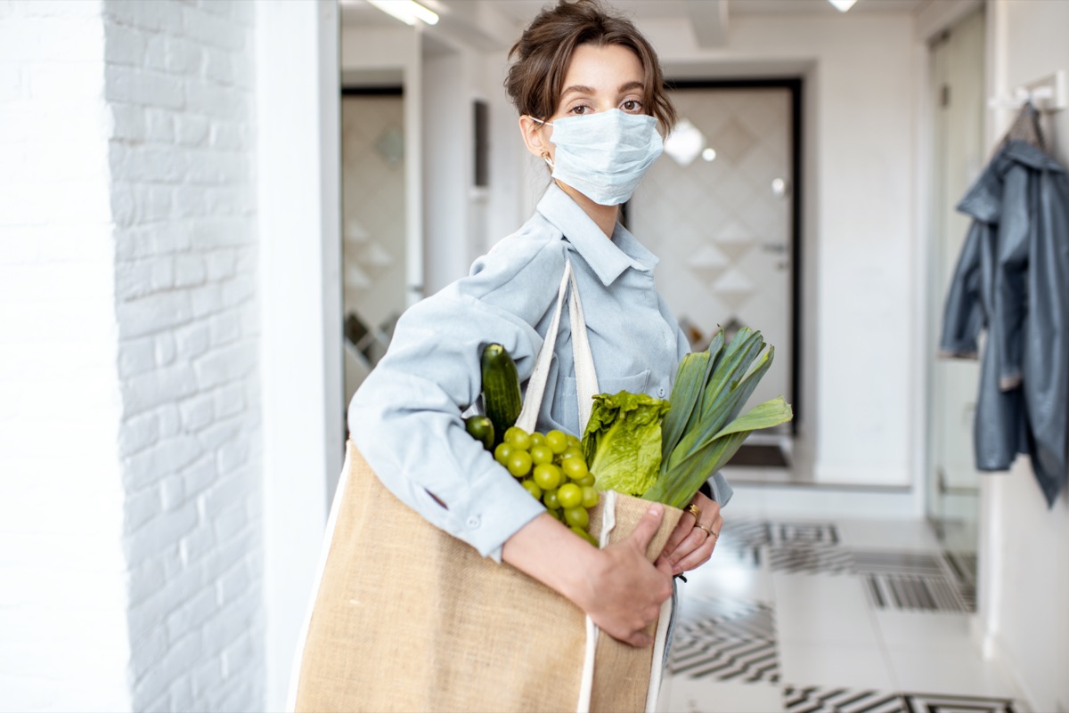 Young woman in medical mask coming home with shopping bag full of fresh food.