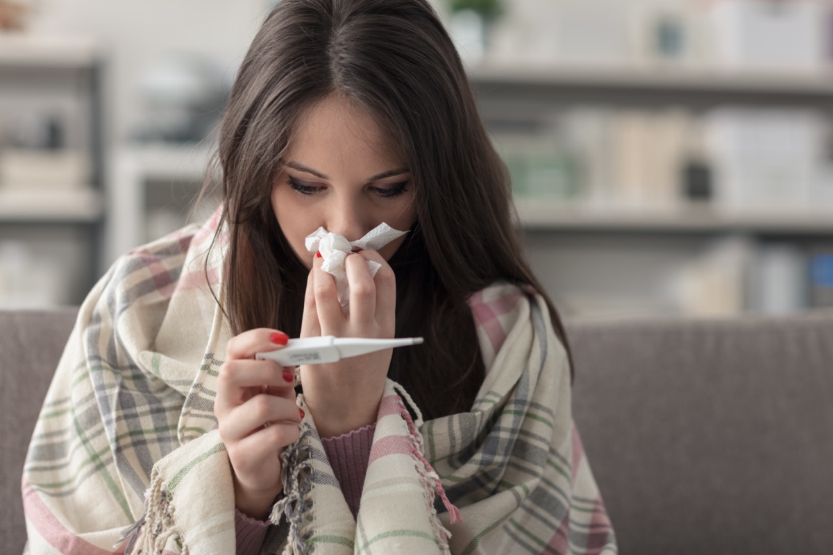 Sick young woman at home on the sofa, she is covering with a blanket, taking temperature and blowing her nose with a tissue