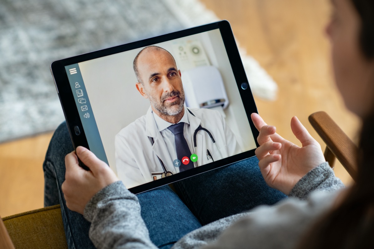 Back view of woman making video call with her doctor while staying at home. Close up of patient in video conferencing with general practitioner on digital tablet. Sick girl in online consultation.