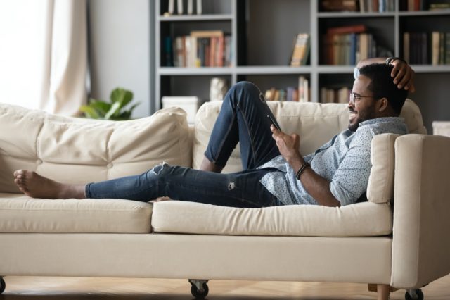 Young black man reclining on couch and reading tablet