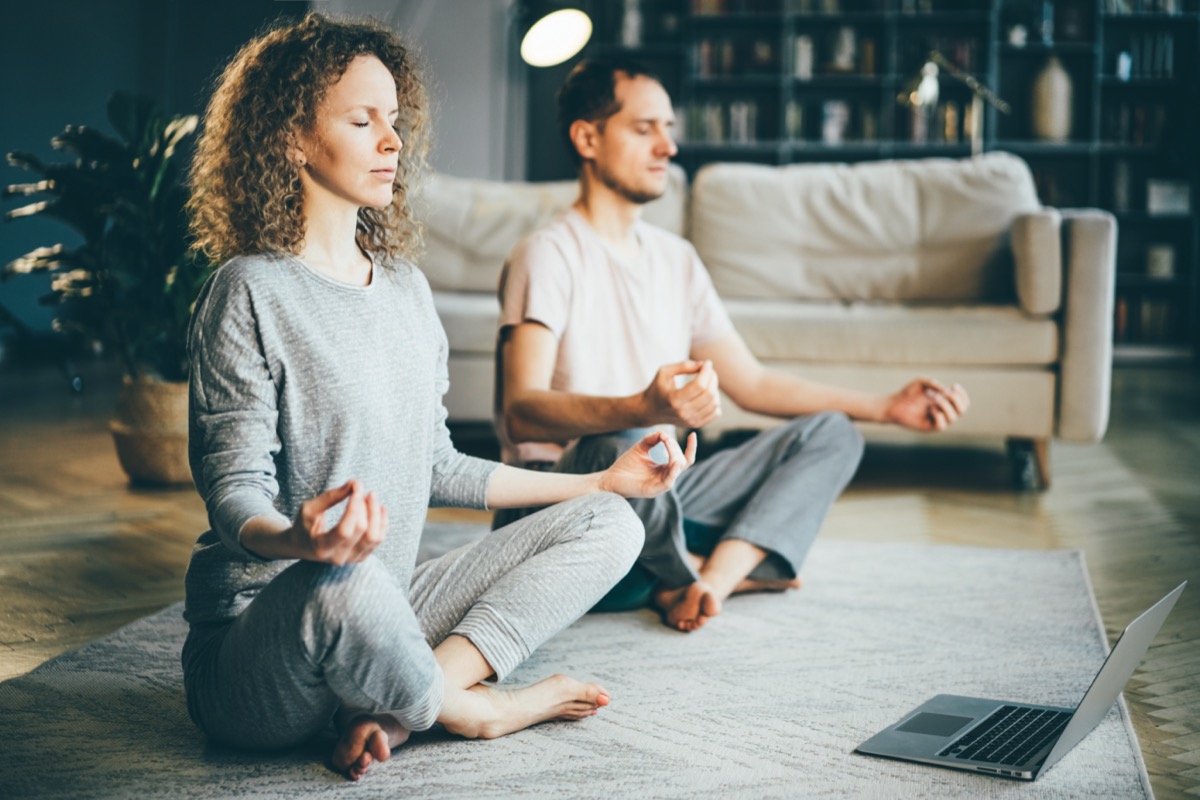 A calm couple in pajamas meditates, listens to spiritual practices on a laptop, sits in a lotus position at home