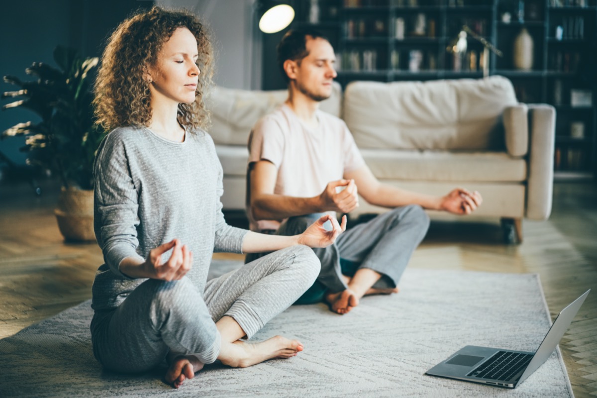 Calm couple in pajamas meditating, listening spiritual practices lessons on laptop, sitting on lotus pose at home