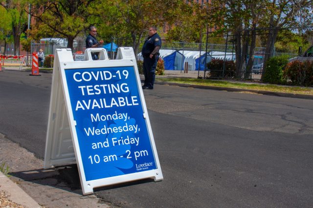 Street sign reads "COVID-19 Testing Available" outside of Lovelace Hospital mobile testing tents guarded by security guards