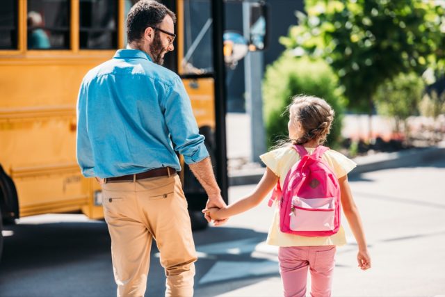 rear view of father and little daughter holding hands and walking to school bus