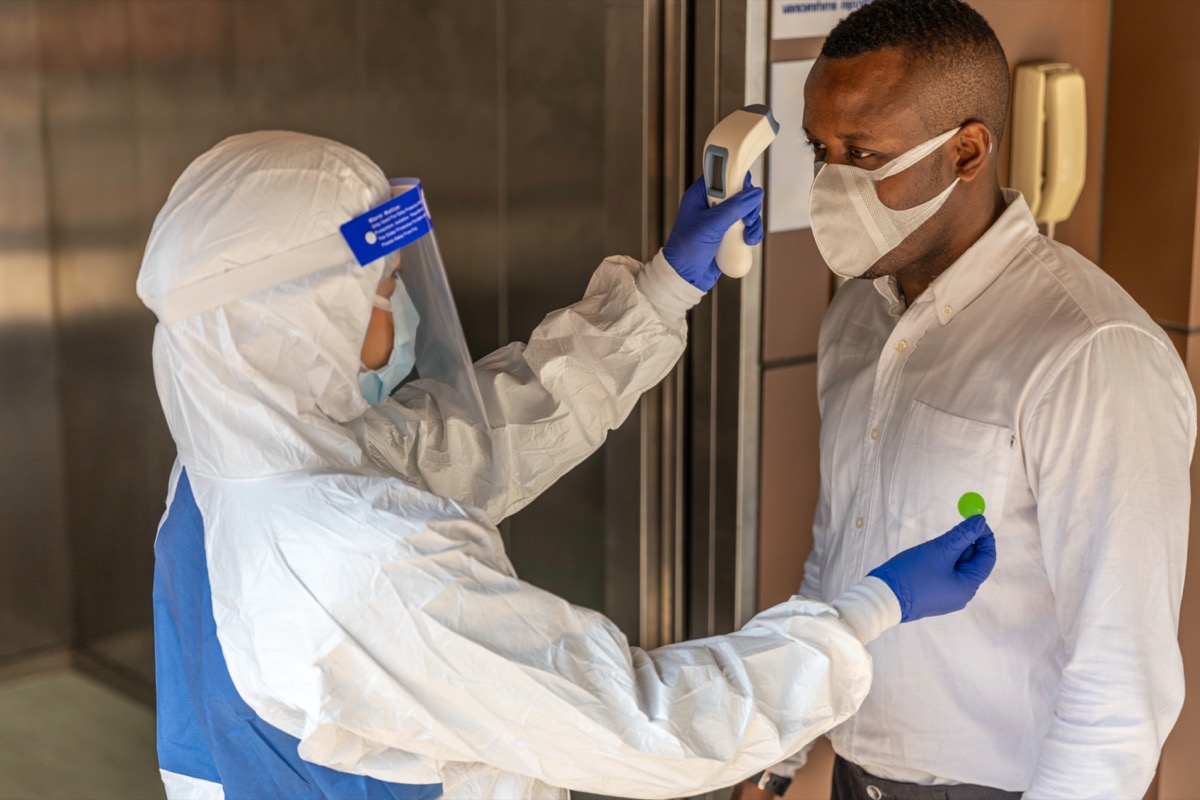 Guard in PPE suit uses infrared thermometer measuring temperature with African male worker scanning for Coronavirus or Covid-19 symptom at office elevator International medical healthcare system