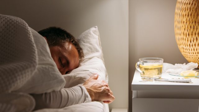 Болен човек спи покрит с одеяло, лежащ в леглото с висока температура, почива в хола.  Той е изтощен и страда от грип с чаша лимонов чай, лекарство.  Сезонът на грипа.