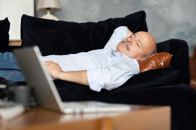 Tired senior hispanic man sleeping on dark blue couch having afternoon nap in living room