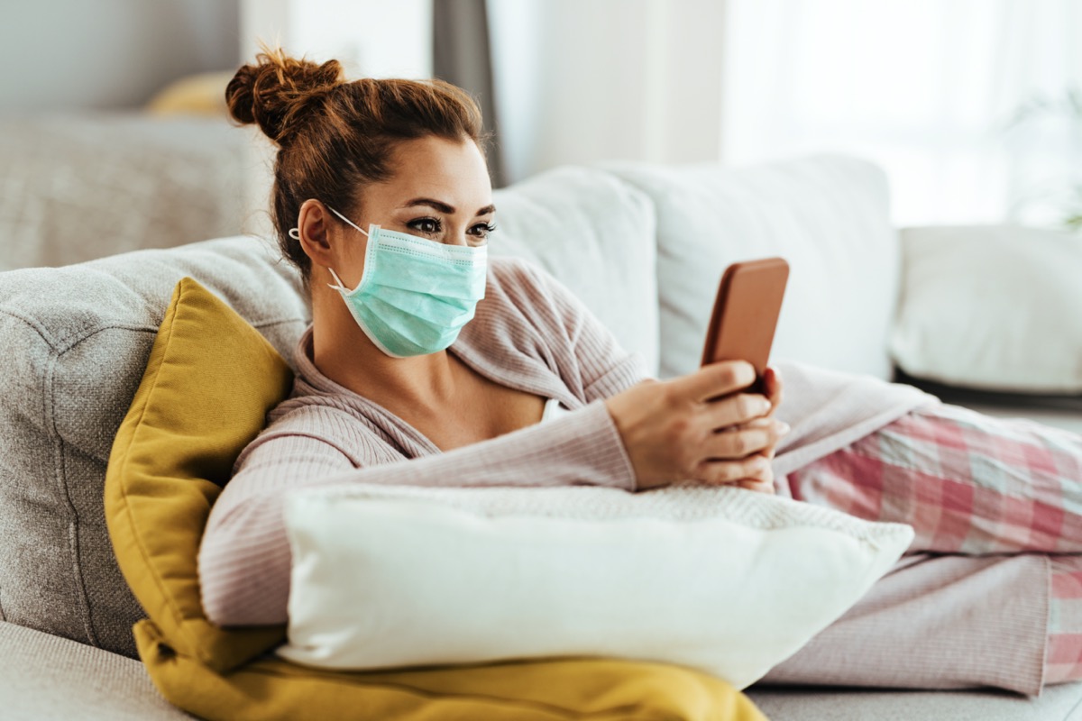 Smiling woman wearing protective face mask while using smart phone and text messaging at home.