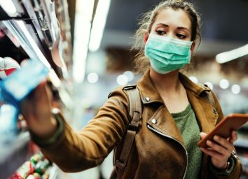 Young woman with face mask using mobile phone and buying groceries in the supermarket during virus pandemic.