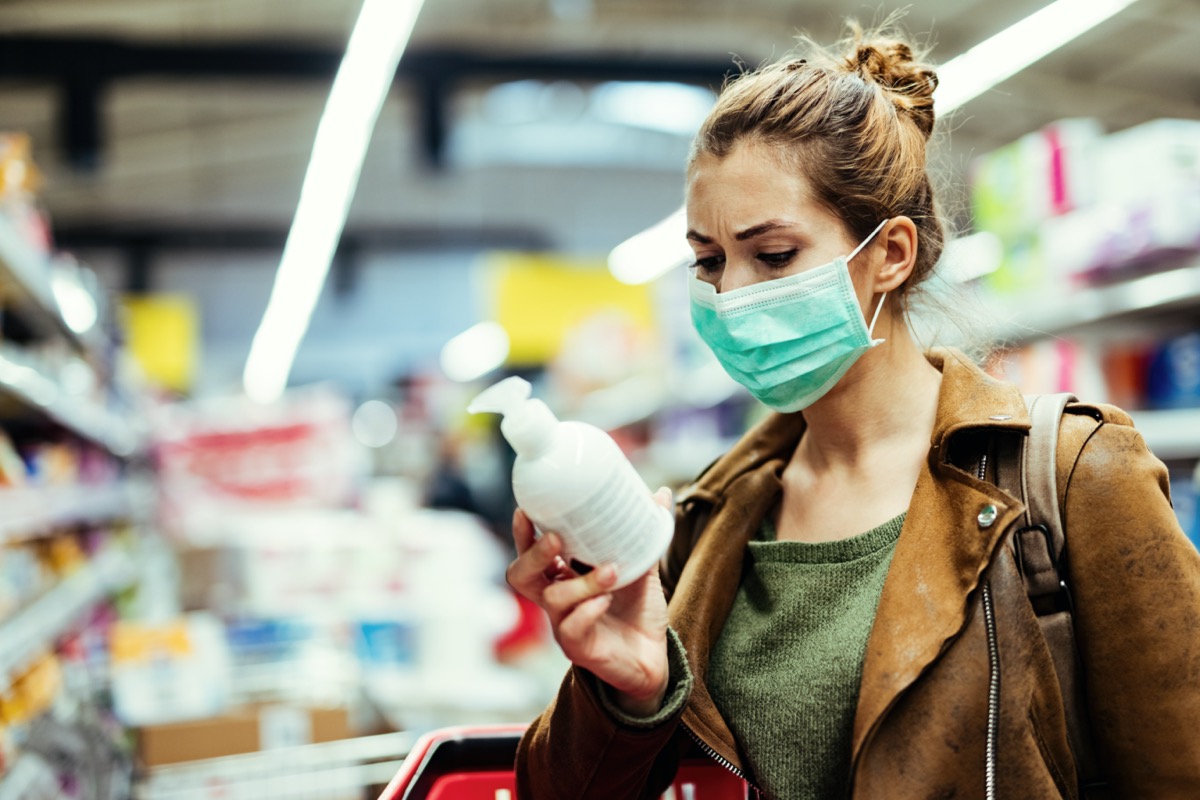 woman with protective mask reading label on the bottle while buying hand soap in the supermarket during virus epidemic