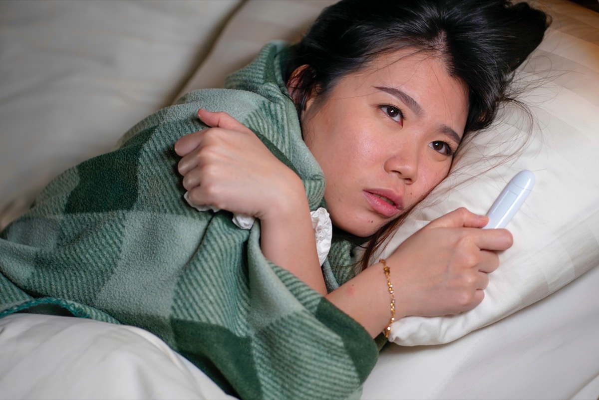 woman lying on bed at home sick suffering cold flu and temperature covered with blanket feeling unwell and feverish