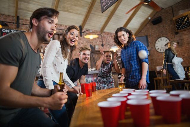 Young friends enjoying beer pong game on table in restaurant
