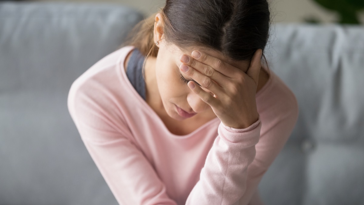 woman sitting on couch in living room at home with closed eyes, holding head with hand, suffering from strong sudden headache or migraine, throbbing pain