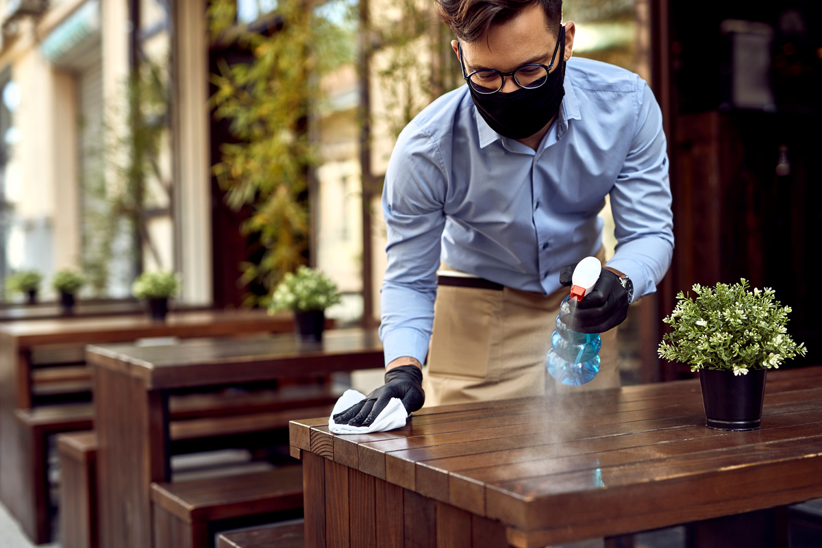 Waiter wearing protective face mask while disinfecting tables at outdoor cafe