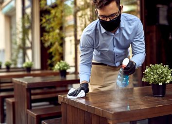Waiter wearing protective face mask while disinfecting tables at outdoor cafe