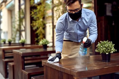 Waiter wearing protective face mask while disinfecting tables at outdoor cafe