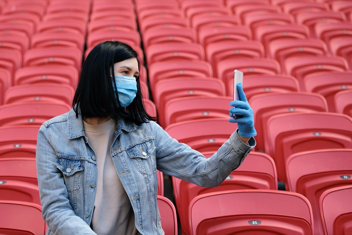 A cheerleader girl in a medical mask and rubber gloves takes a selfie or photographs on a smartphone alone in an empty stadium with red seats