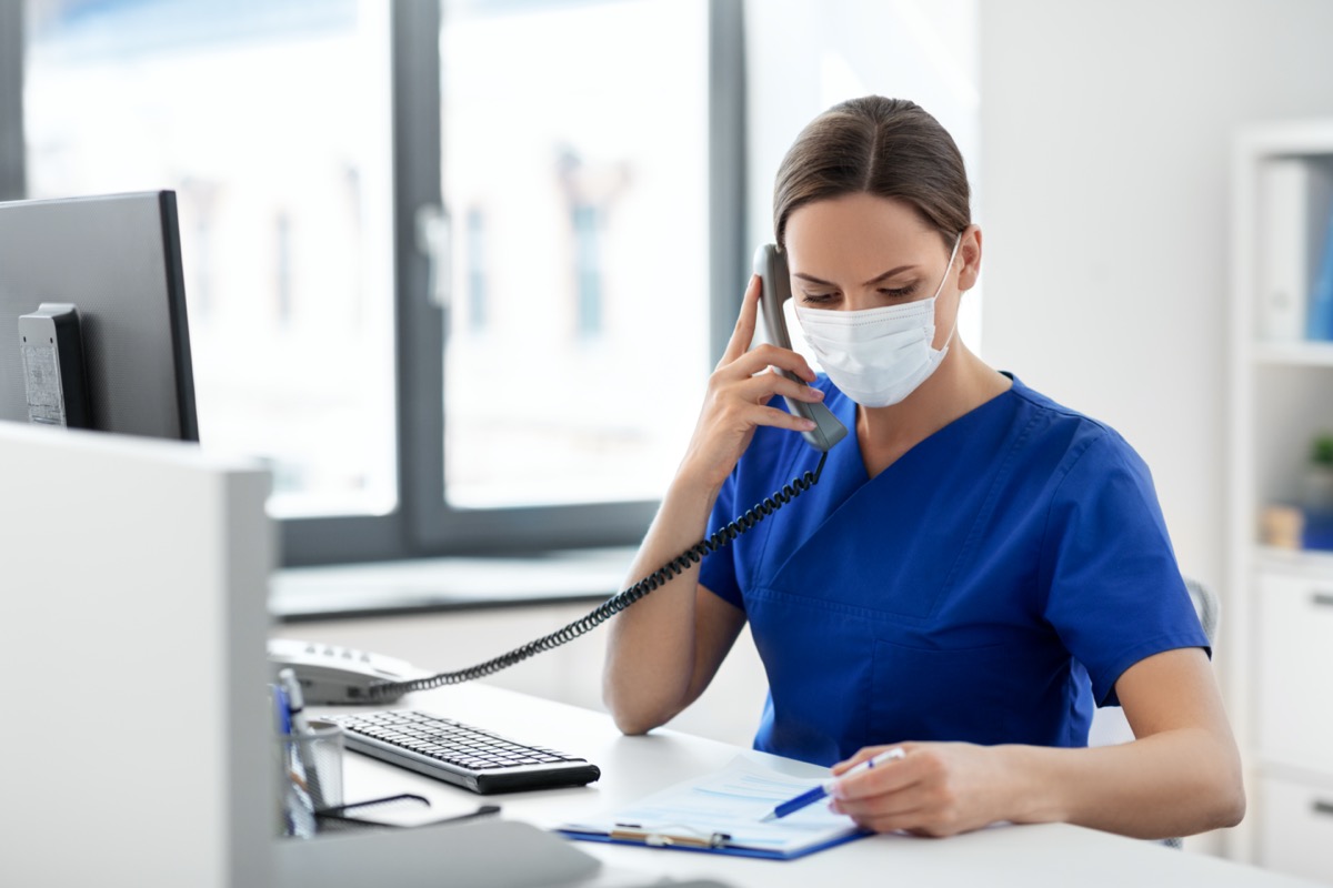 female doctor or nurse wearing face protective medical mask for protection from virus disease with computer and clipboard calling on phone at hospital