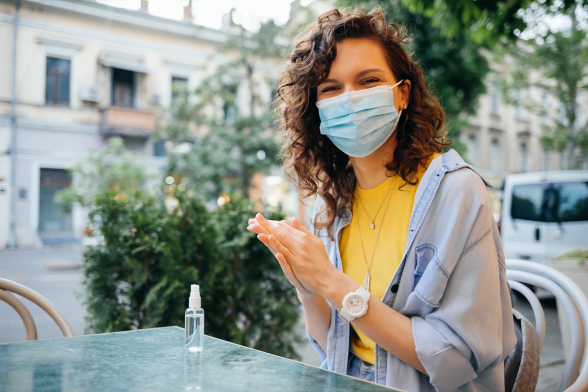 Happy young woman wearing protective face mask disinfects her hands with alcohol sanitizer while sitting at table in restaurant on summer day.