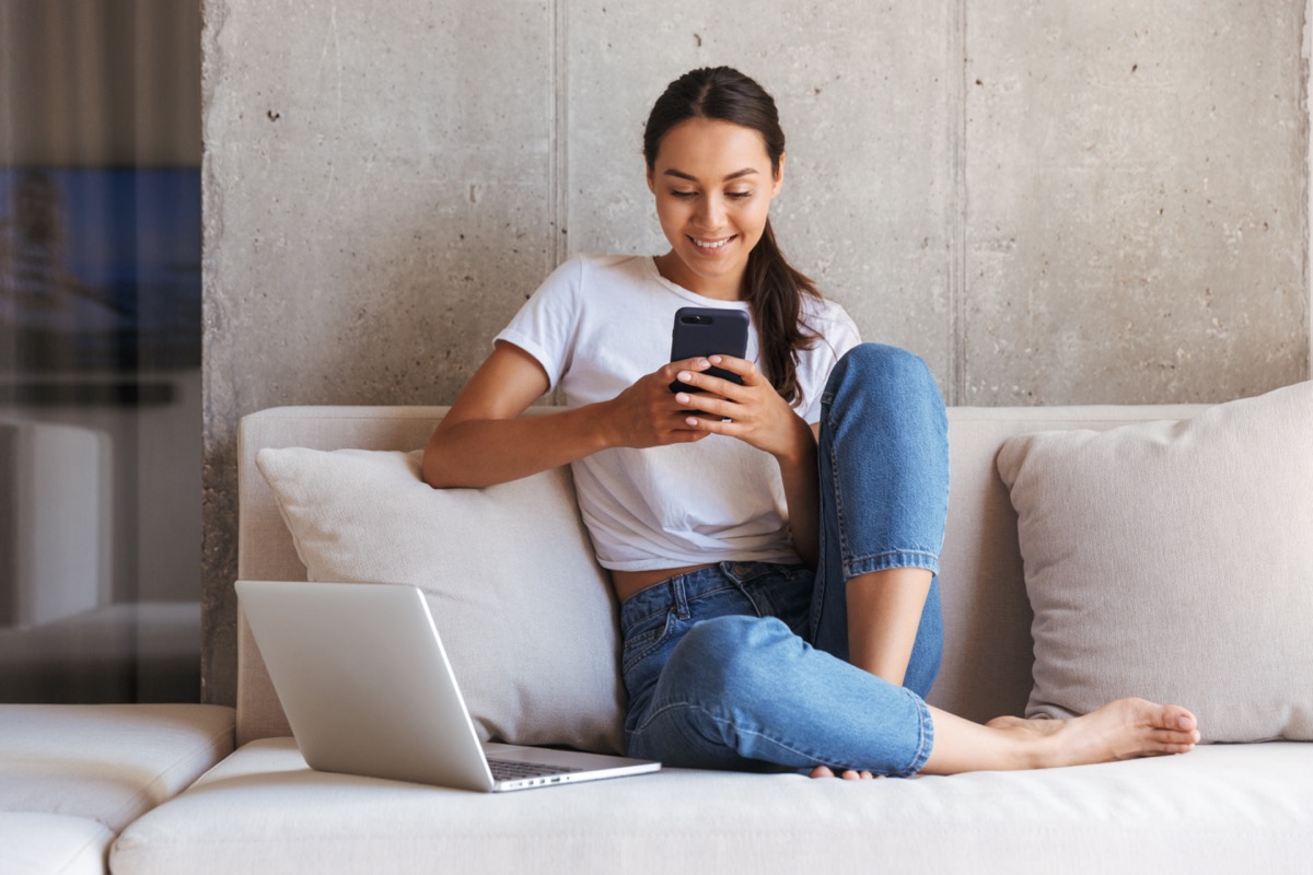 woman using mobile phone while sitting a couch at home with laptop computer.