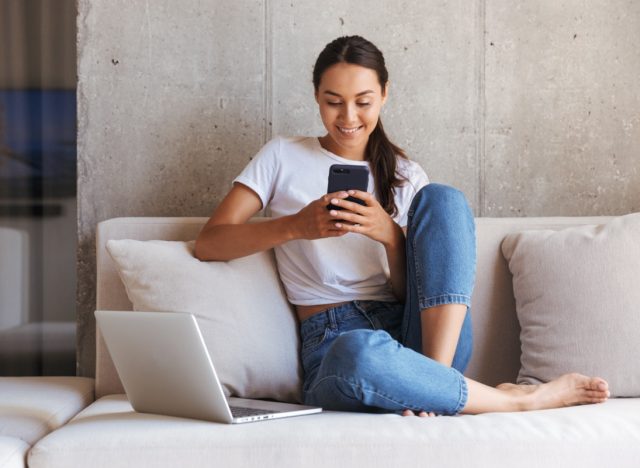 woman using mobile phone while sitting a couch at home with laptop computer.