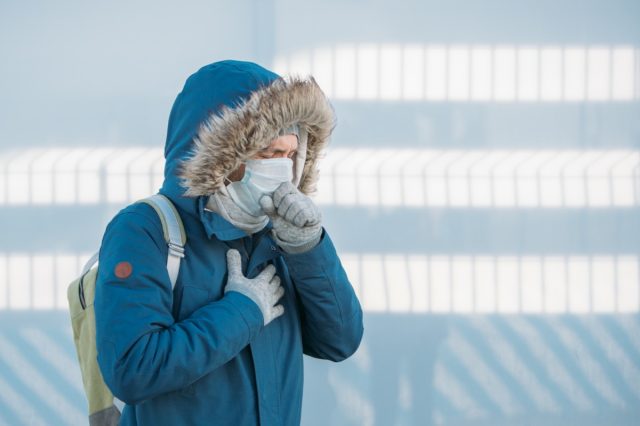 Portrait of sick young man in blue jacket put on a hood, having a cold, feeling unwell, coughing, wearing medical face mask, outdoors