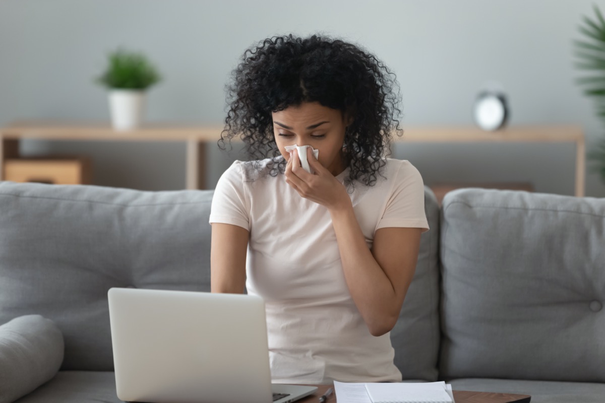 Allergic african woman blowing nose in tissue sit on sofa at home office study work on laptop