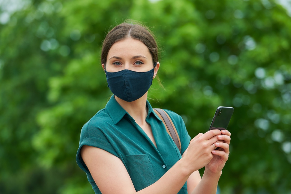 A close portrait of a woman in a medical face mask is holding a smartphone while walking in the park.