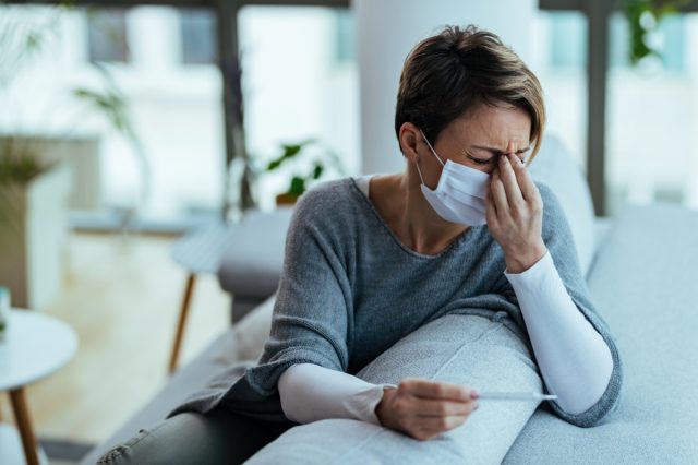 woman with face mask feeling sick and holding her head in pain while measuring temperature at home.