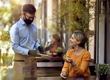 Happy waiter wearing protective face mask while showing menu on digital tablet to female guest in a cafe.