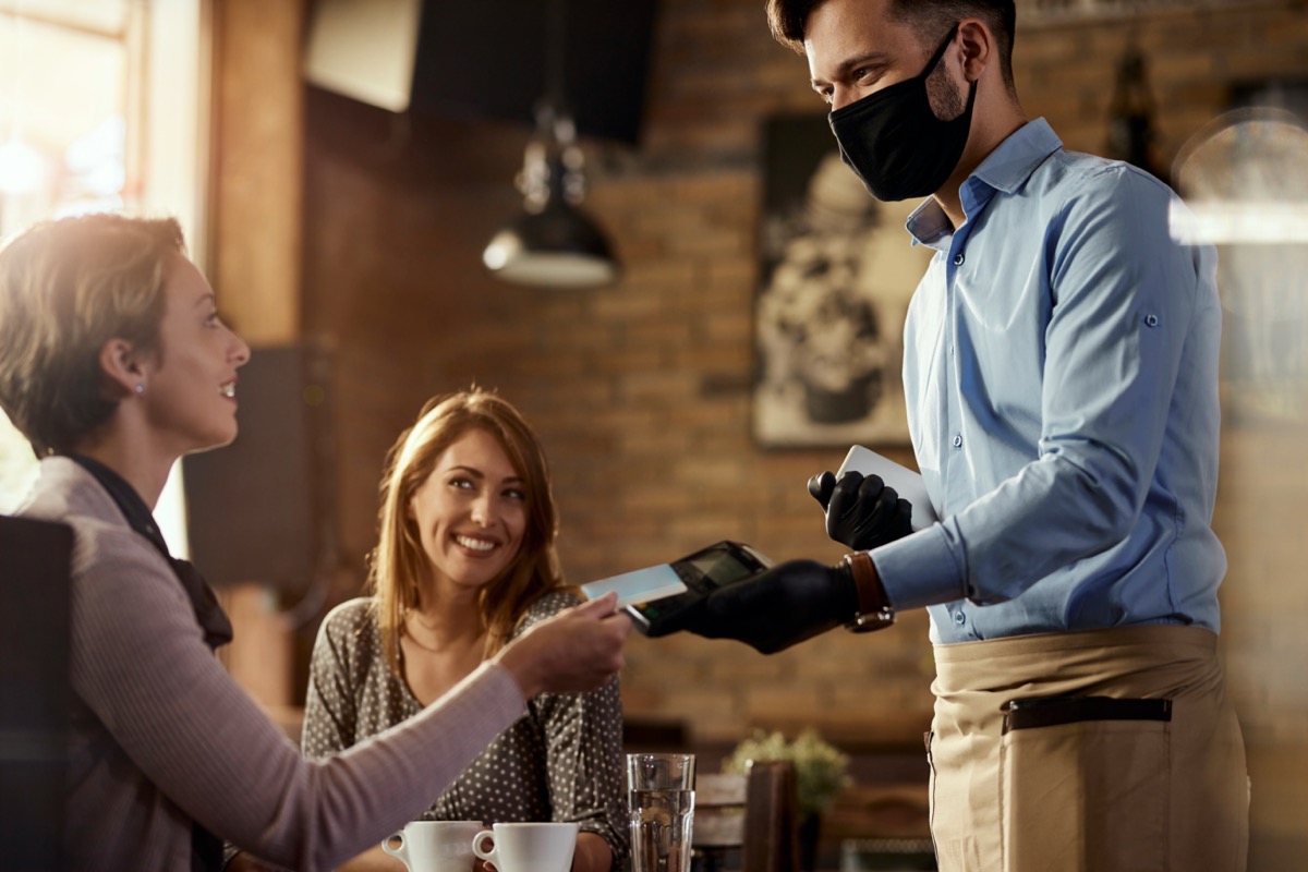 Young waiter wearing protective face mask while his guests are making contactless payment with credit card in a cafe.