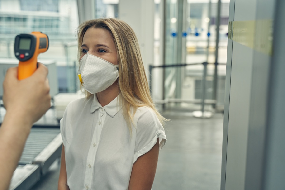 female wearing protective mask while standing opposite the worker of airport and checking temperature