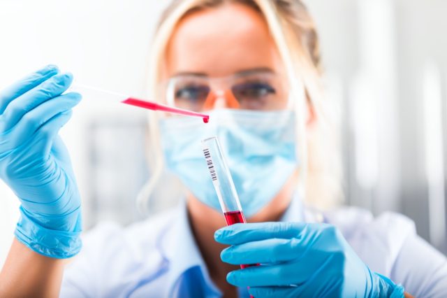 Young attractive concentrated female scientist in protective eyeglasses, mask and gloves dropping a red liquid substance into the test tube with a pipette in the scientific chemical laboratory