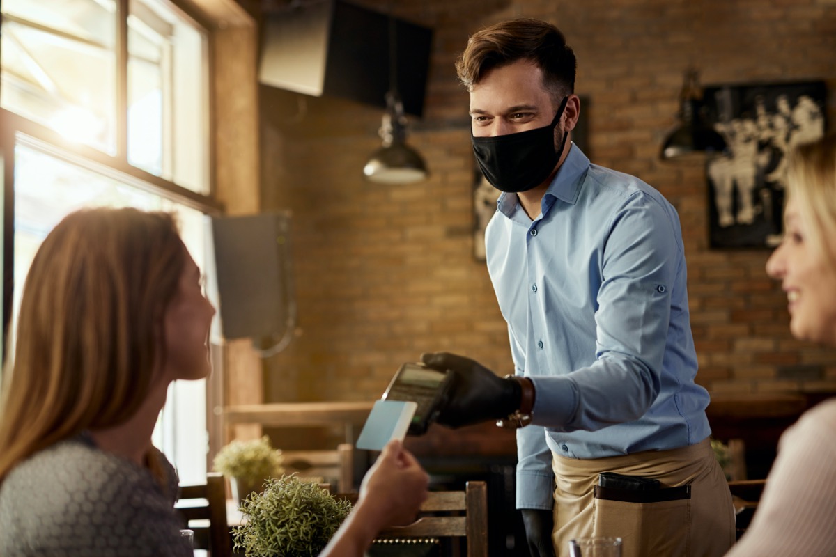 Female customer making contactless payment to a waiter who is wearing protective face mask in a cafe.