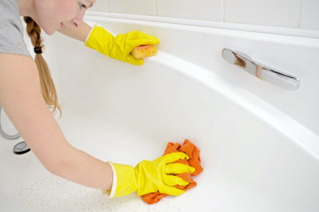 Female washing bathtub in yellow rubber gloves with orange sponge.