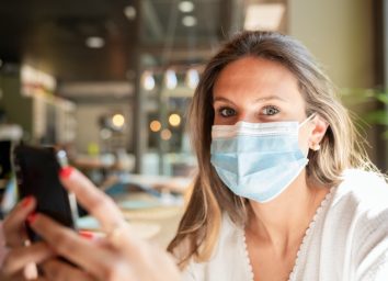Woman in a face mask sitting at a bar, looking at the camera while holding her smart phone in her hands