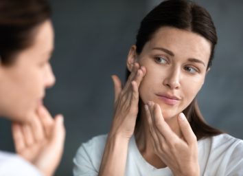 woman looking at red acne spots on chin in mirror, upset young female dissatisfied by unhealthy skin