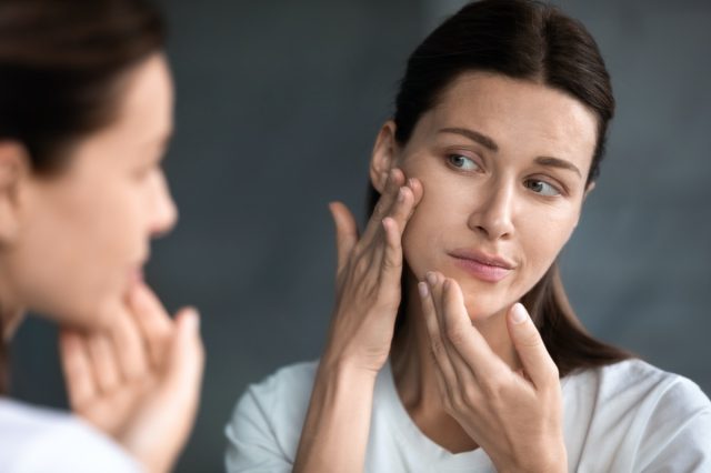 woman looking at red acne spots on chin in mirror, upset young female dissatisfied by unhealthy skin