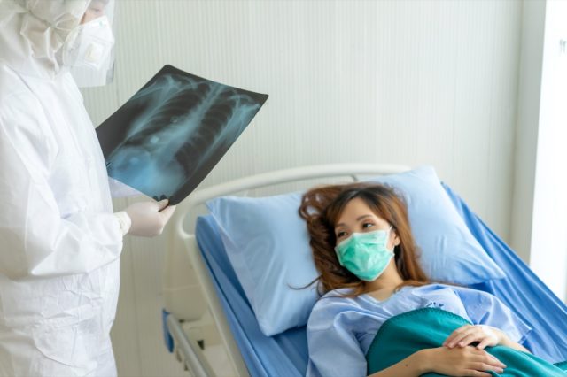 Patienten, die mit Maske auf einem Krankenhausbett liegen und einen Lungenröntgenfilm betrachten, während der Arzt das Ergebnis liest und die Behandlung empfiehlt