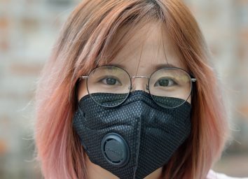 woman with dyed hair and eyeglasses wears a black protective face mask with breathing valve.