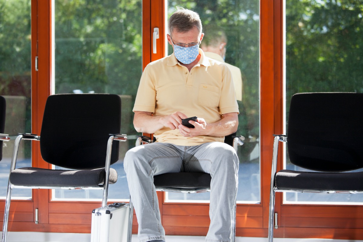Mature man with face mask sitting in a bright waiting room of a hospital or an office looking at smart phone